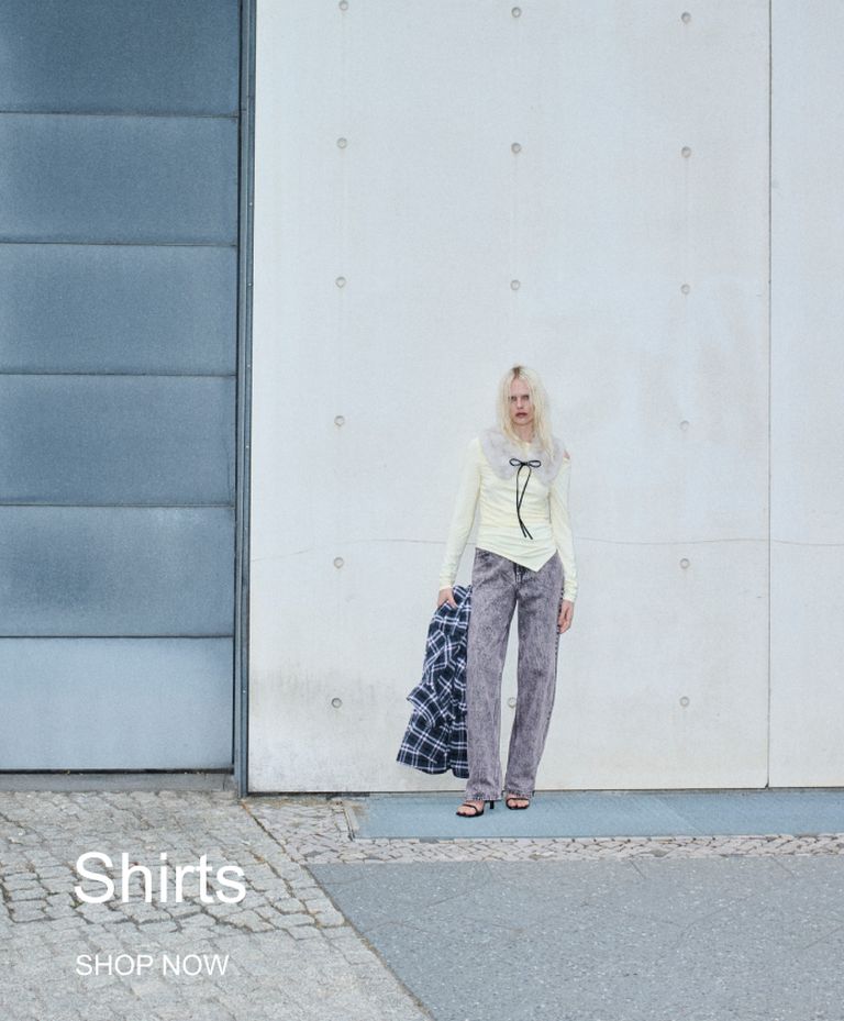 Person in a light top, holding a plaid shirt, stands against a concrete wall with "Shirts" and "Shop Now" text on the side.