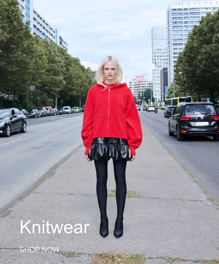 A person in a red hoodie and black skirt stands on a city sidewalk, flanked by cars. "Knitwear" and "Shop Now" text at the bottom.