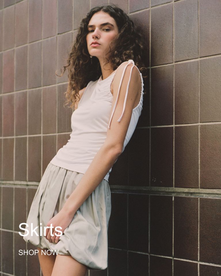A person with curly hair wearing a sleeveless top and a light-colored skirt leans against a tiled wall. Text reads "Skirts" and "Shop Now."