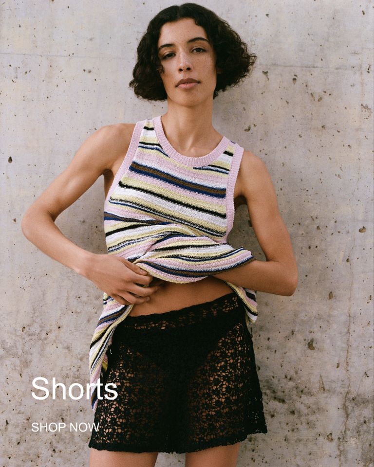 Person wearing a striped tank top and black lace shorts, standing against a concrete wall. Text reads "Shorts, Shop Now."