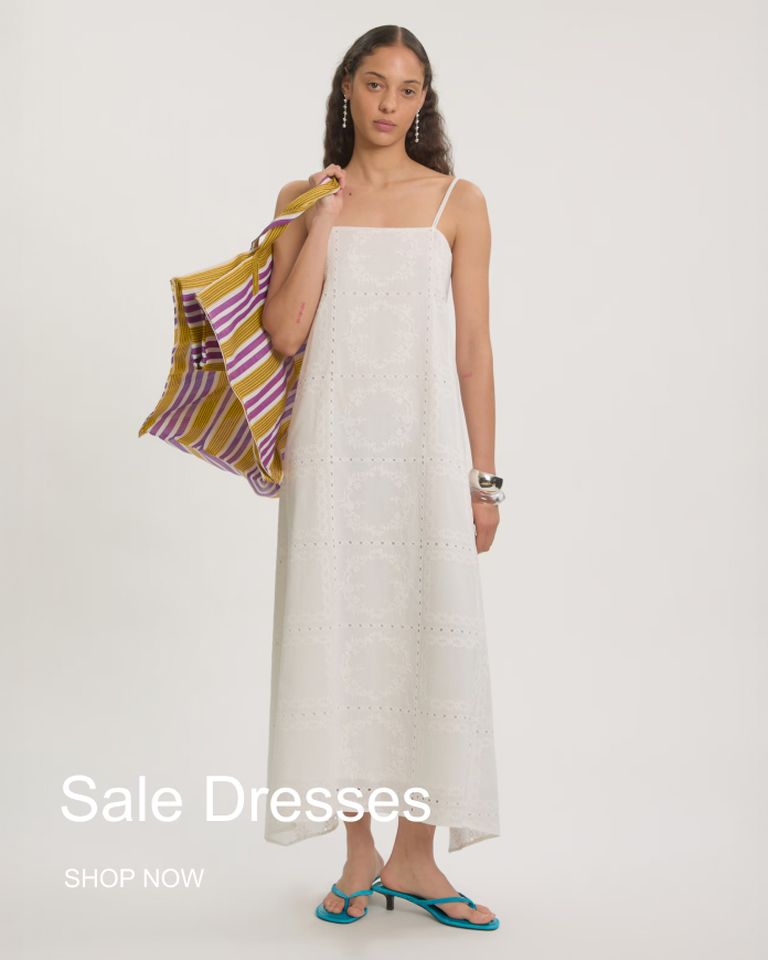 Woman in a white summer dress holding a striped bag, wearing blue sandals. Text reads "Sale Dresses, Shop Now."