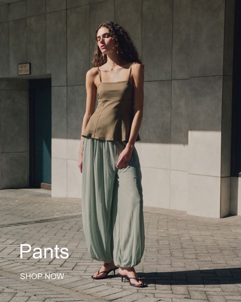 A woman wearing a sleeveless top and flowy pants stands in sunlight against a tiled wall. Text reads "Pants" and "Shop Now."