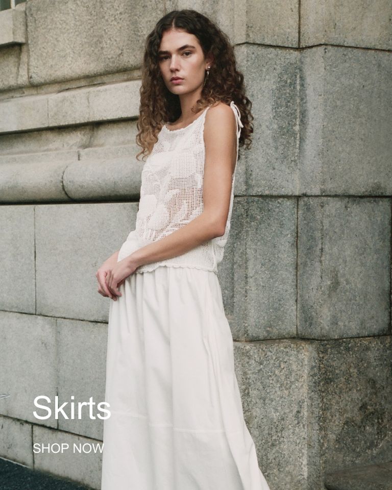 A person wearing a sleeveless knit top and a long white skirt stands against a stone wall. Text reads "Skirts SHOP NOW."
