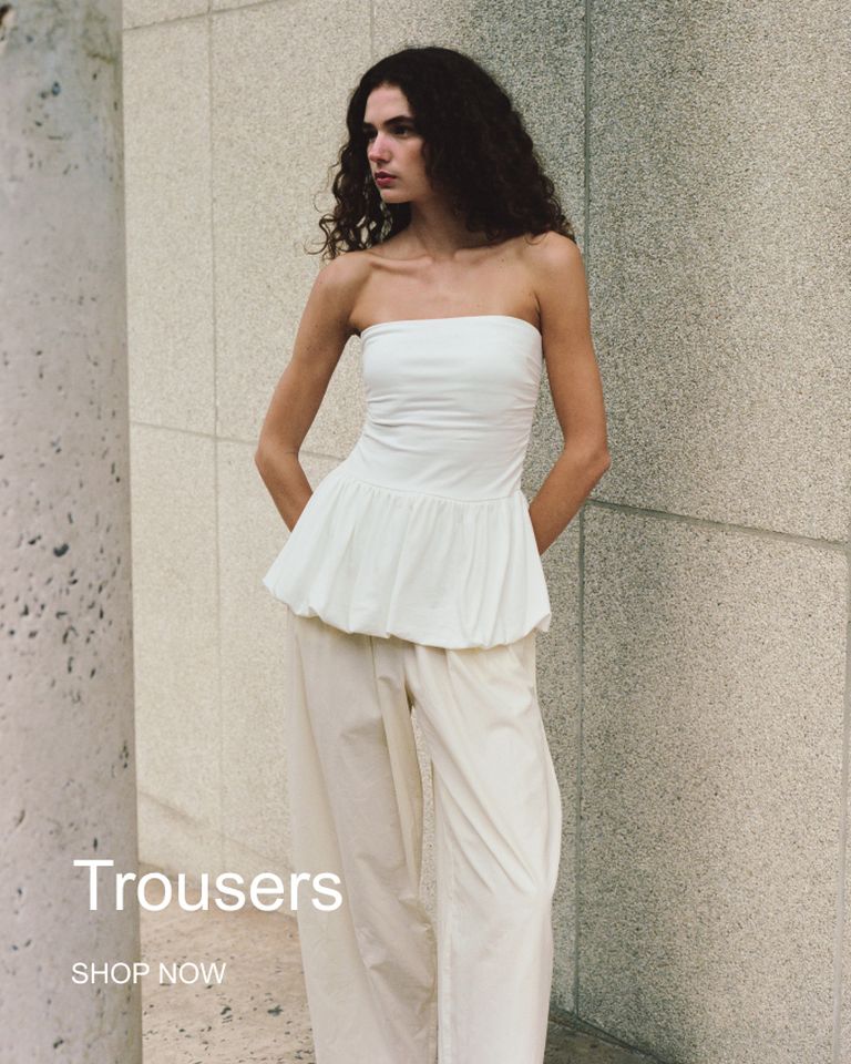 Person in a strapless white top and loose beige trousers, standing against a textured wall. Text: "Trousers" and "Shop Now."