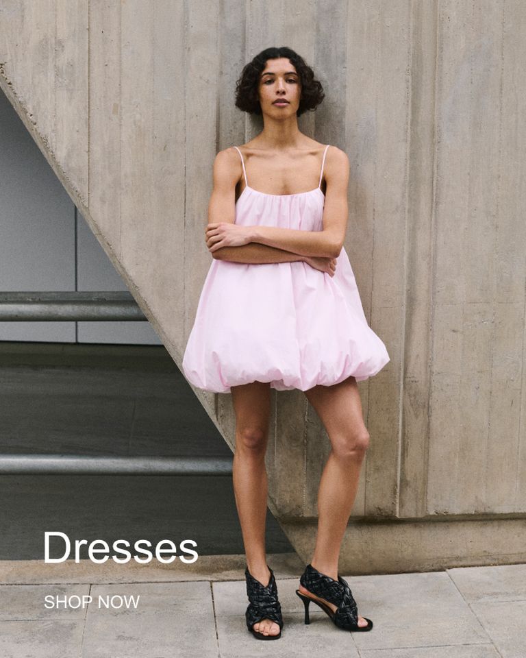 Person in a light pink dress and black heels stands against a textured concrete wall. Text reads: "Dresses. Shop Now."