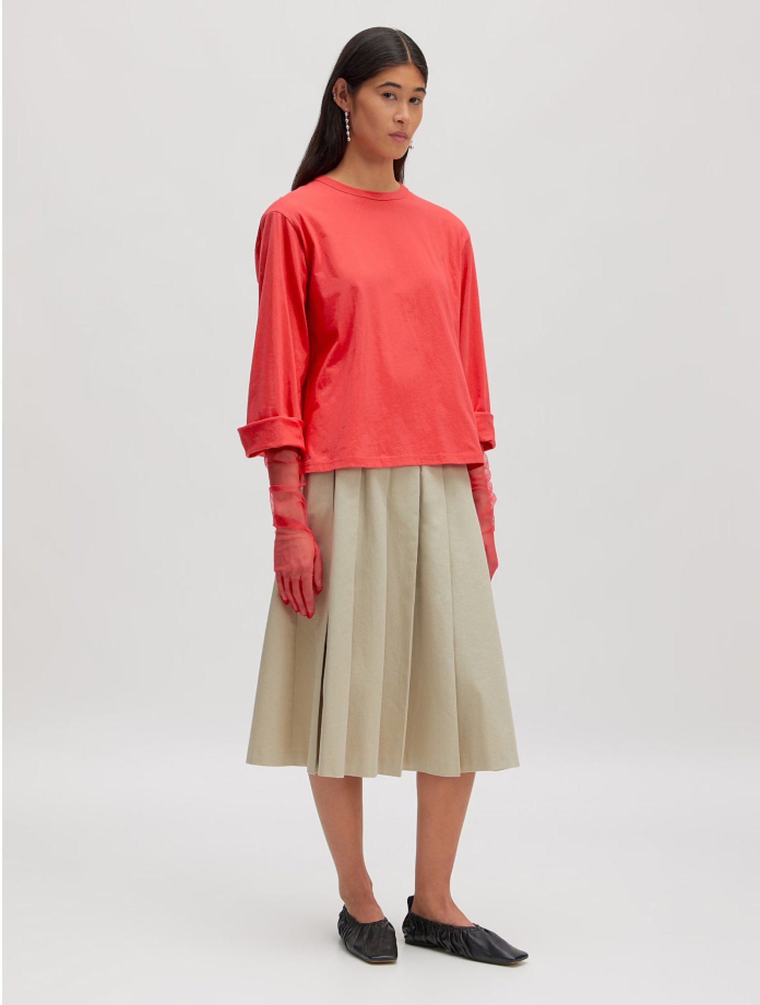 Woman wearing a red long-sleeve shirt, beige pleated skirt, red gloves, and black slip-on shoes, standing against a plain background.
