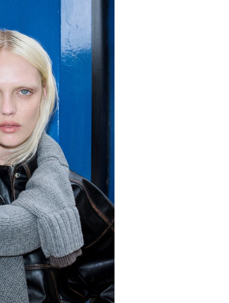 Person with blonde hair wearing a gray sweater and black leather jacket, standing against a blue background.