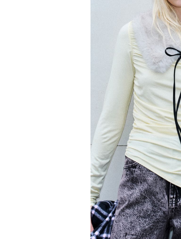 Person in a light yellow fur-collar top with long sleeves and gray jeans. A black ribbon hangs from the collar.
