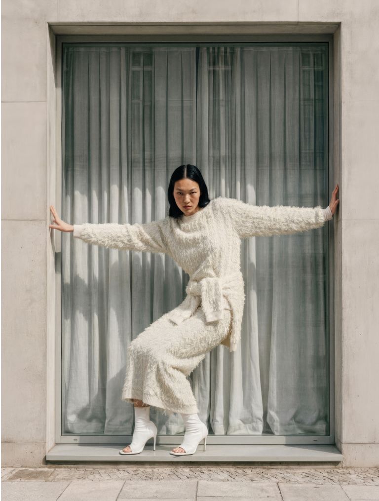 Woman in a textured white outfit and high heels poses confidently in a window frame with curtains, arms spread to the sides.