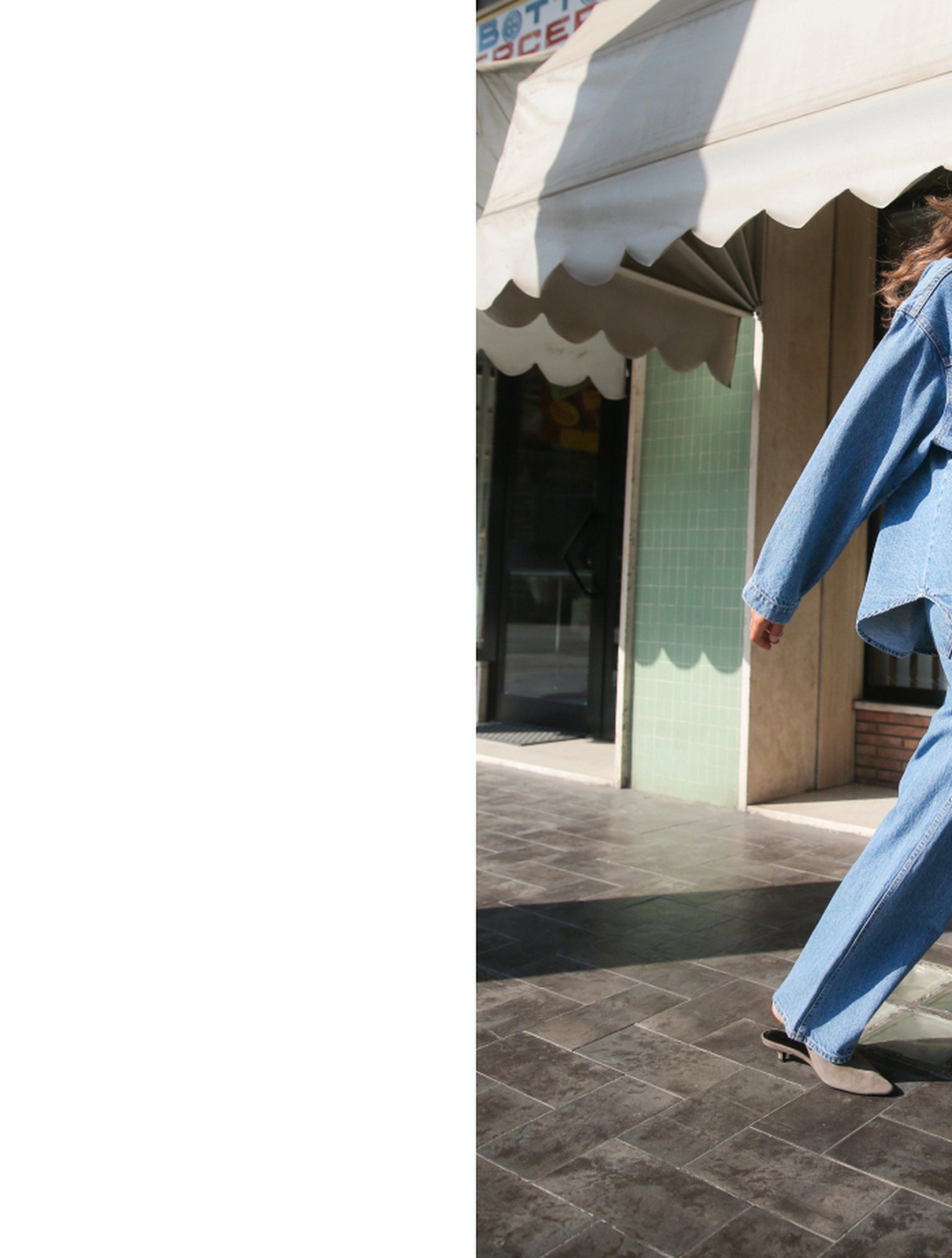 Person in a blue denim outfit walking on a sunlit sidewalk near a building with green tiles and a white awning.