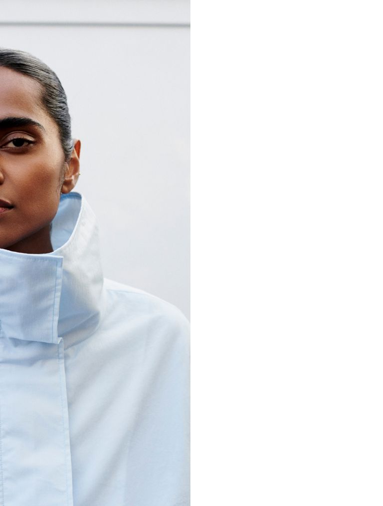 Person wearing a high-collared white coat, with a minimalist background and half of their face visible.