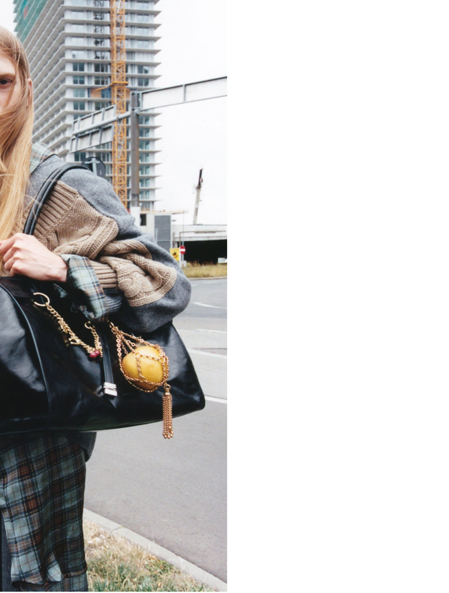 Person holding a large black handbag with a gold charm, wearing a plaid coat and beige sweater, standing near a construction site.