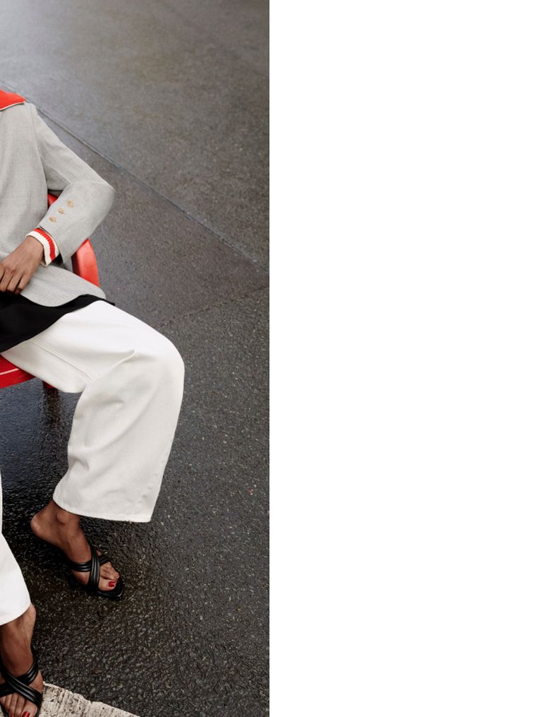 Person seated on a red chair, wearing a gray blazer, white pants, and black sandals, against a dark floor background with a white wall on the right.