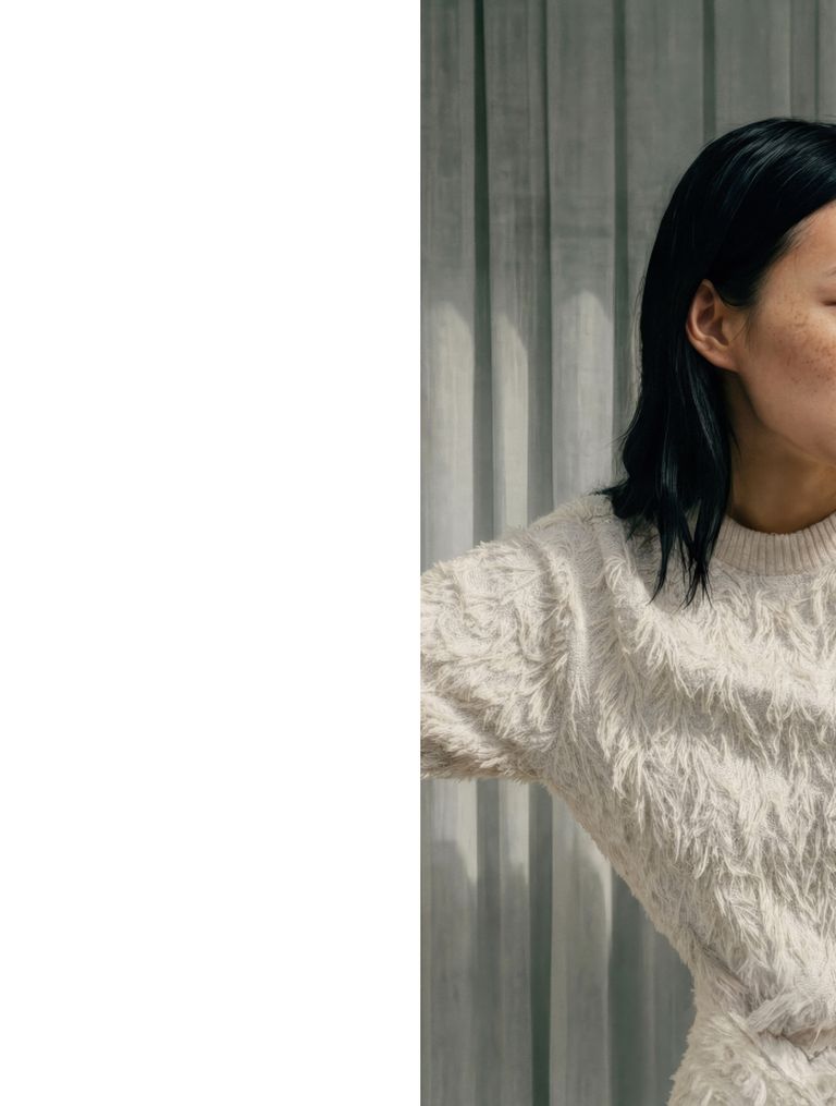 Person in a textured cream sweater stands against sheer curtains, facing to the right.