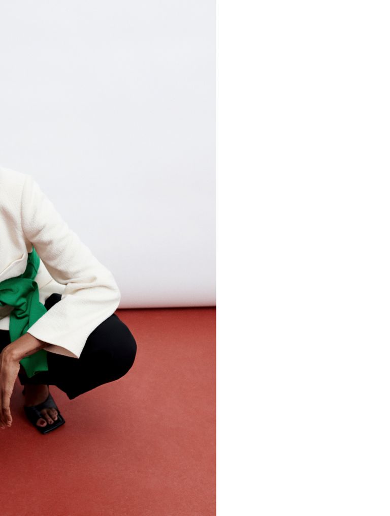Person crouching on a red floor wearing a white blazer, green top, and black pants. Their hand rests on their knee against a white backdrop.