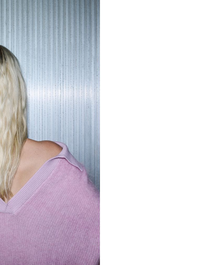 The image shows the back of a person with long, wavy blonde hair and a pink sweater against a textured, vertical striped background.