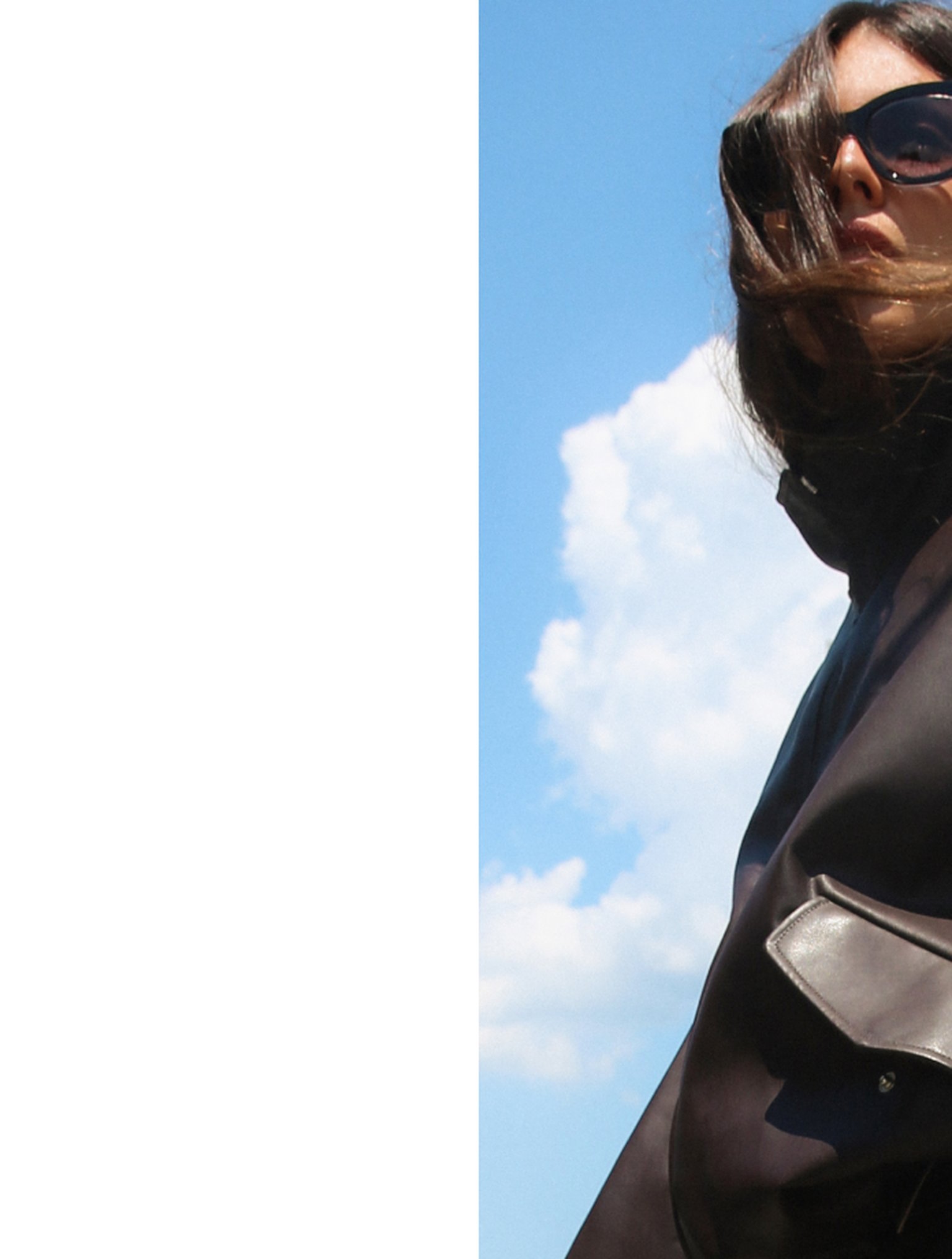 Woman in sunglasses with windblown hair, wearing a dark coat, stands against a blue sky with clouds.