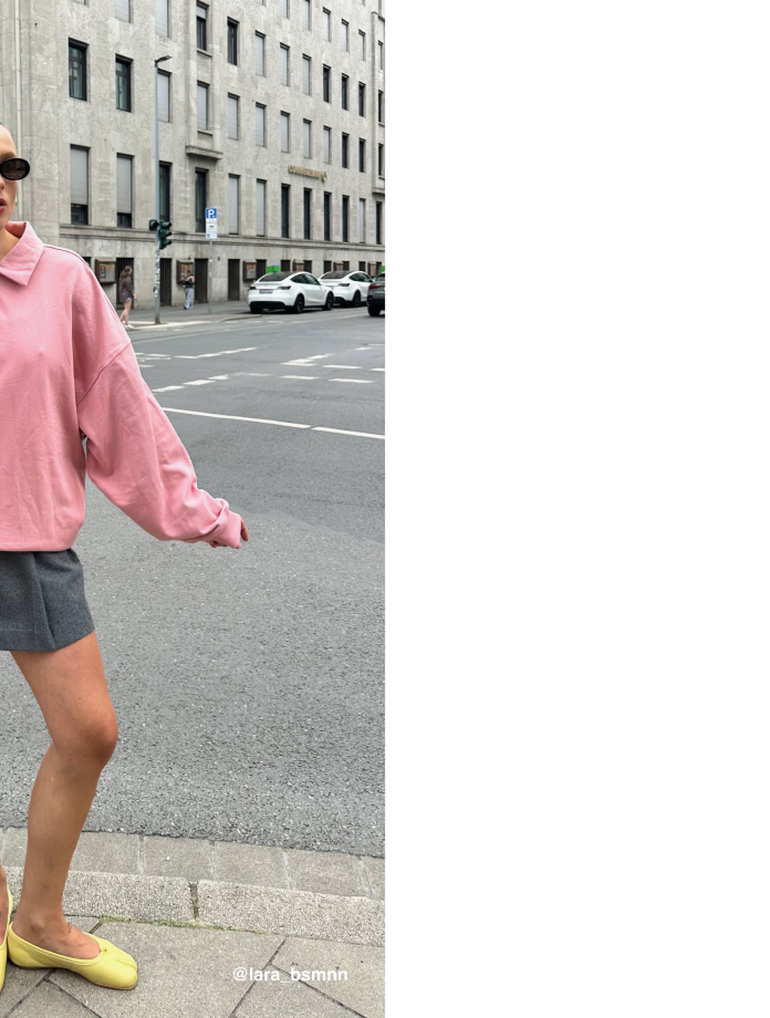 Person in a pink sweater and gray shorts stands on a city street corner, wearing yellow shoes and sunglasses.