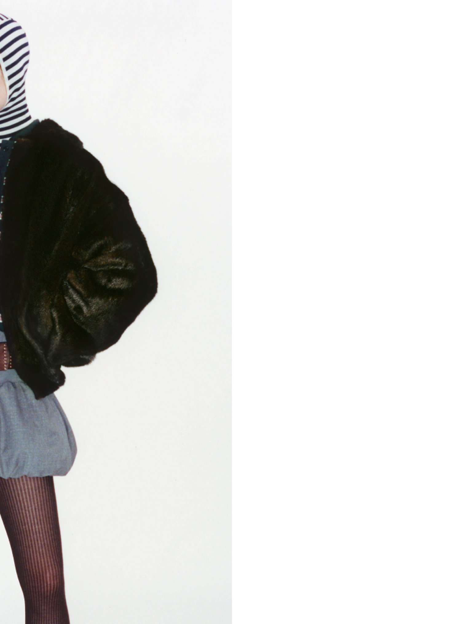 Person wearing a striped top, black fur jacket, and gray skirt against a white background.