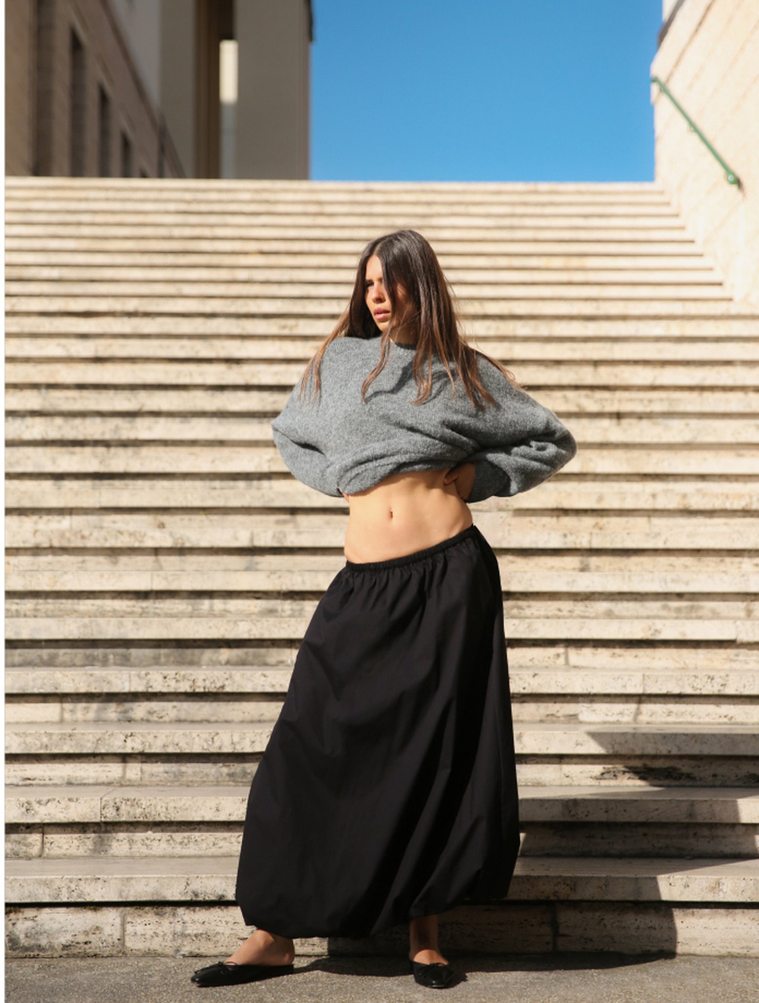 Woman with long hair, wearing a gray sweater and black skirt, stands on stone steps under a clear blue sky.