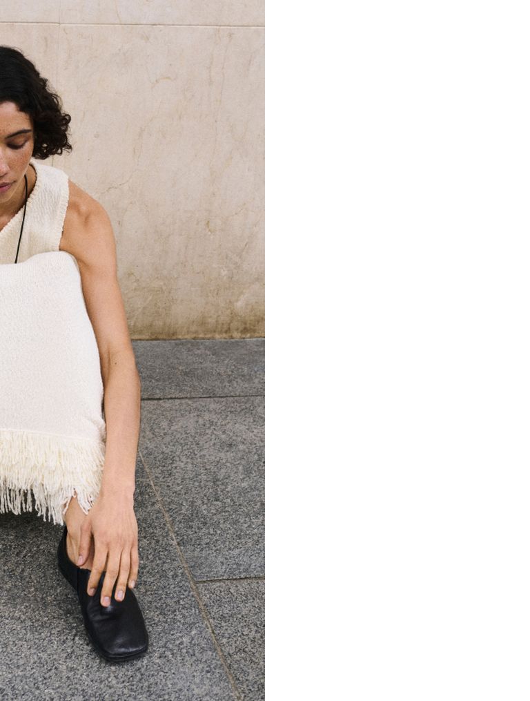 Person in a white fringed dress crouching on a stone floor, head partially visible, leaning against a beige wall.