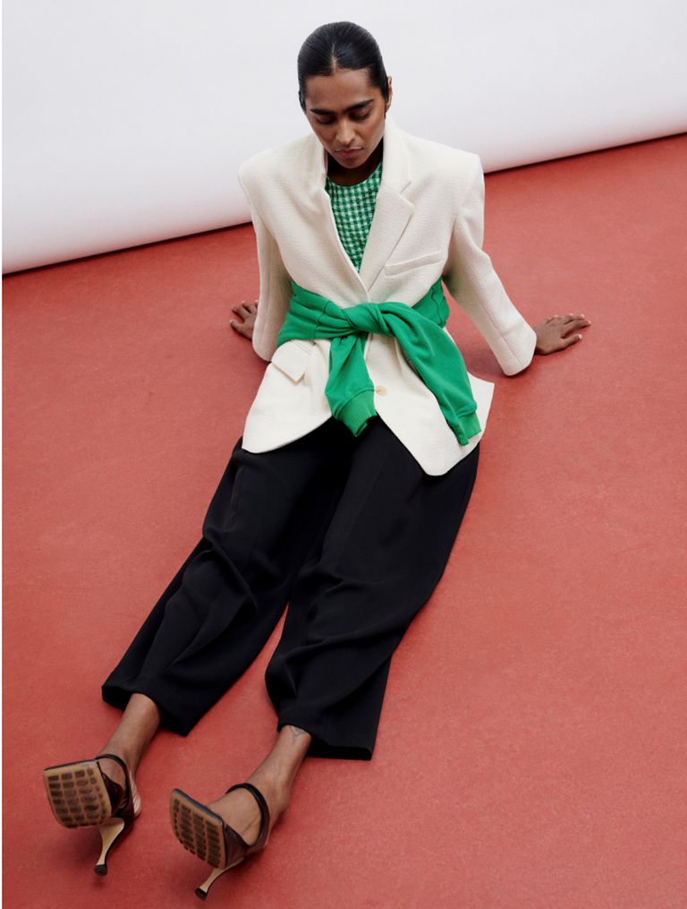 Person sitting on a red floor, wearing a white blazer with a green sweater tied at the waist, green checkered shirt, black pants, and heels.