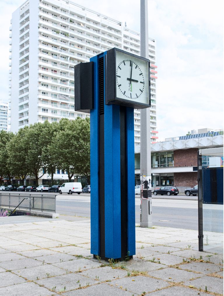 A modern blue clock tower stands on a city street, with a tall building and trees in the background. Cars are parked along the street.
