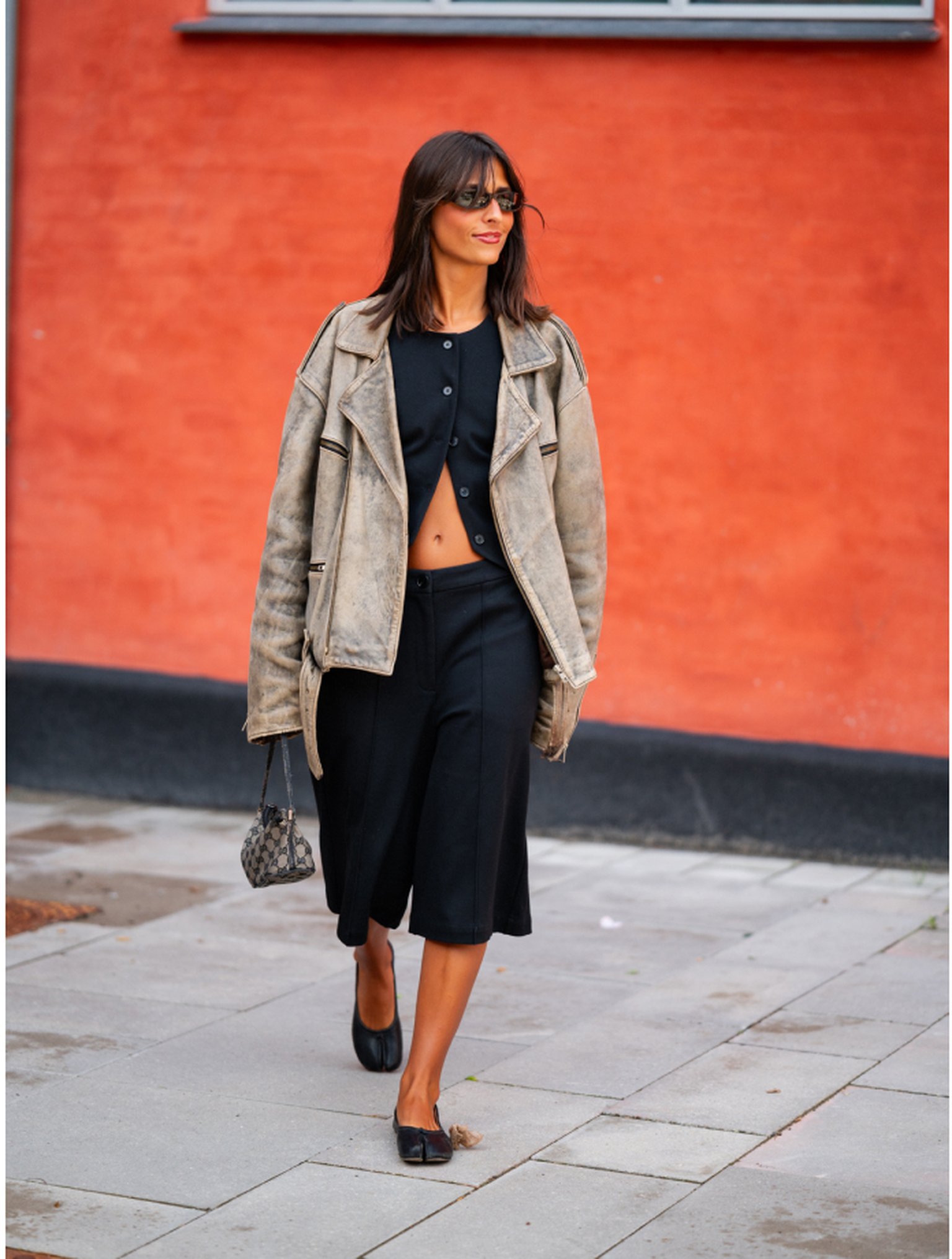 Woman in a black outfit and oversized beige jacket walks on a sidewalk against a red wall, carrying a patterned handbag and wearing sunglasses.
