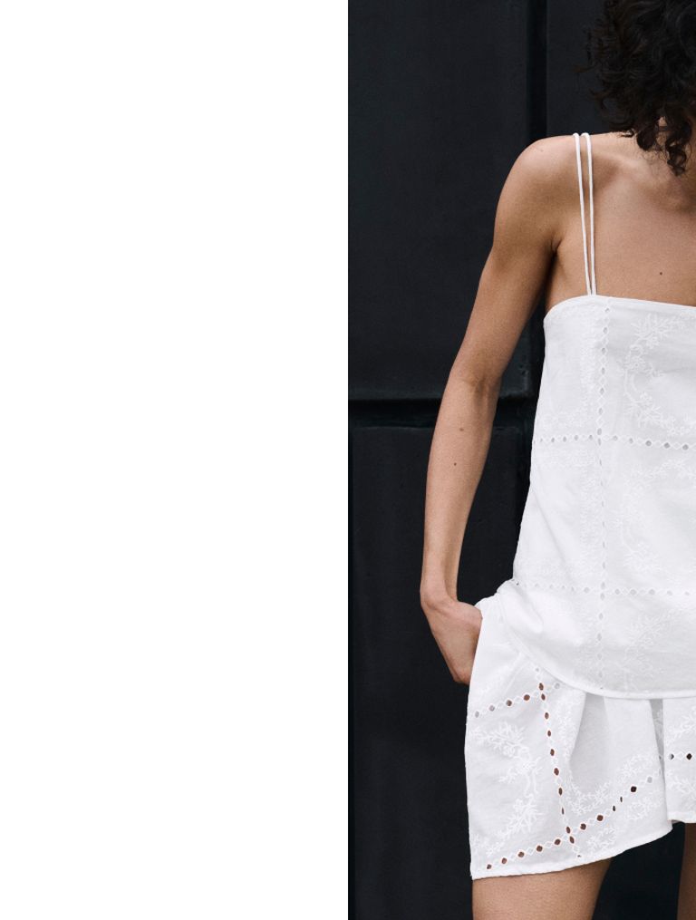 Person in a white, strappy dress with embroidered details stands against a dark background.