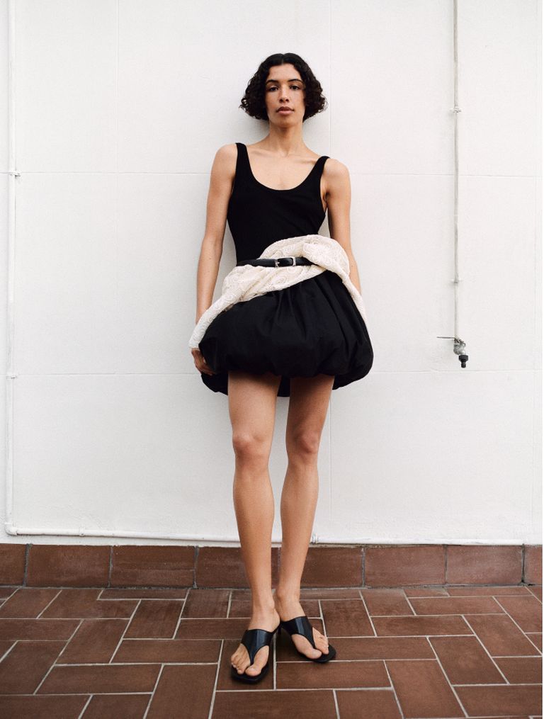 Person in a black outfit with a voluminous skirt and white wrap stands against a white wall, wearing black sandals.