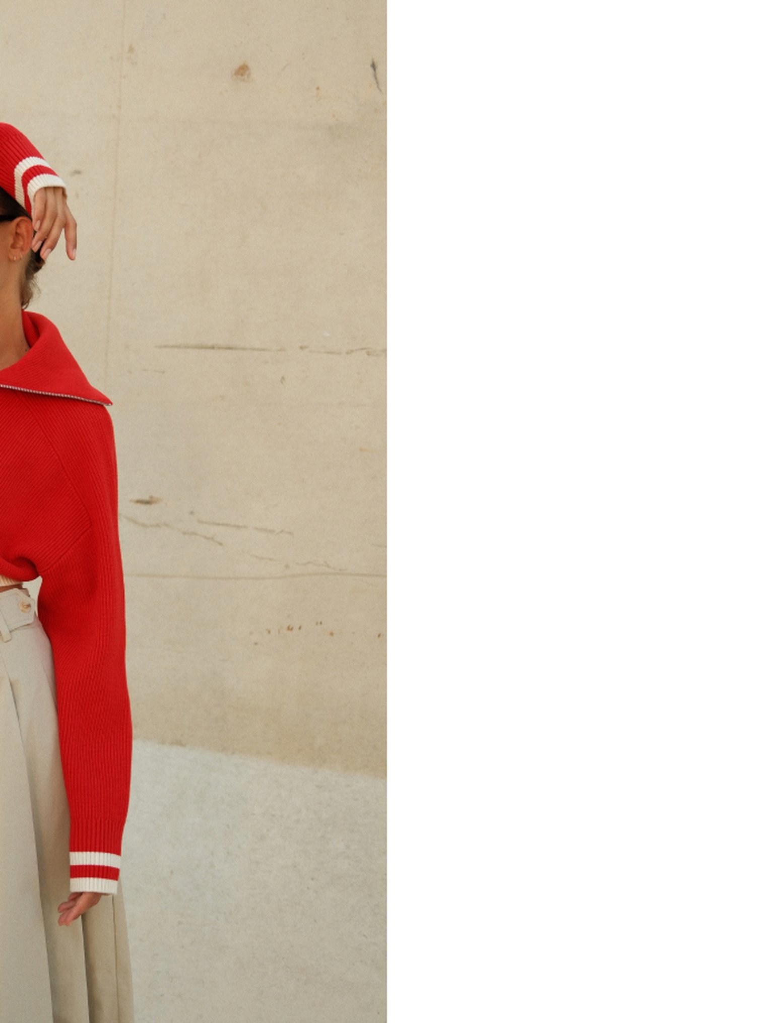 Person in a red sweater and light trousers stands against a beige wall. Their face is partially visible, with the focus on clothing details.