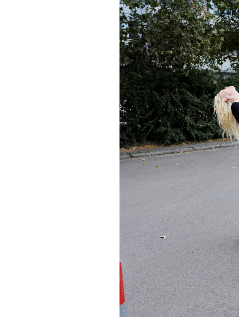 A person with long blonde hair flips their hair back on a paved surface near green bushes.