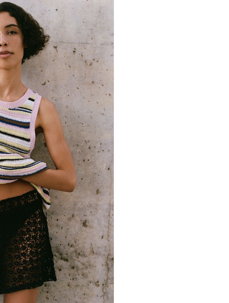 Person in a striped, sleeveless top and black lace skirt standing against a textured wall.
