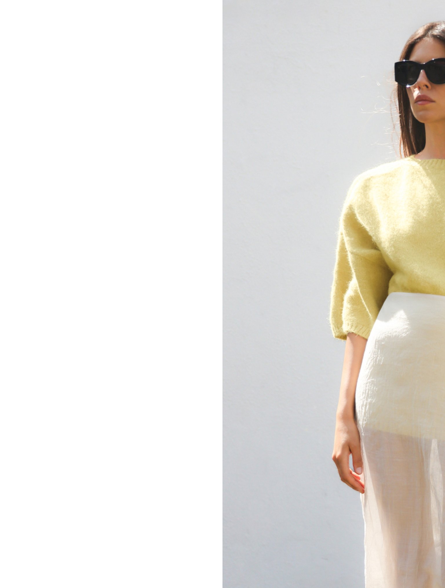 Woman in a yellow sweater and sheer skirt, wearing sunglasses, stands against a plain white wall, partially visible.