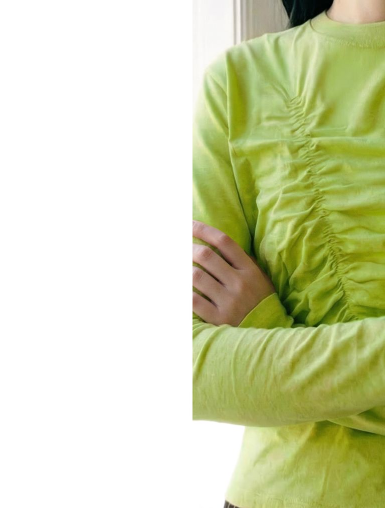 Person wearing a light green, ruched long-sleeve shirt with arms crossed, standing near a light-colored wall.