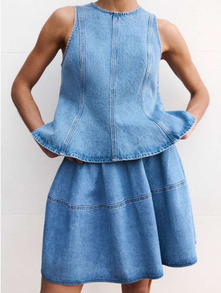 Person wearing a sleeveless denim peplum top and matching skirt, hands in pockets, against a plain background.