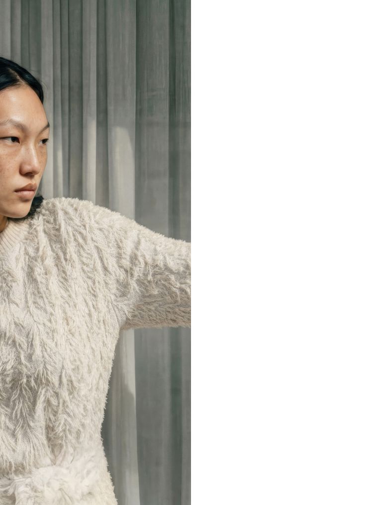 Person in a textured white sweater stands in front of sheer gray curtains, looking to the left with a neutral expression.