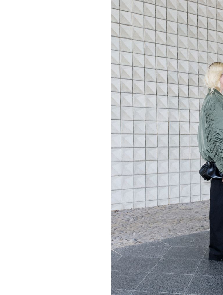 A person with blonde hair, wearing a green jacket and holding a black bag, stands near a tiled wall with a geometric pattern.