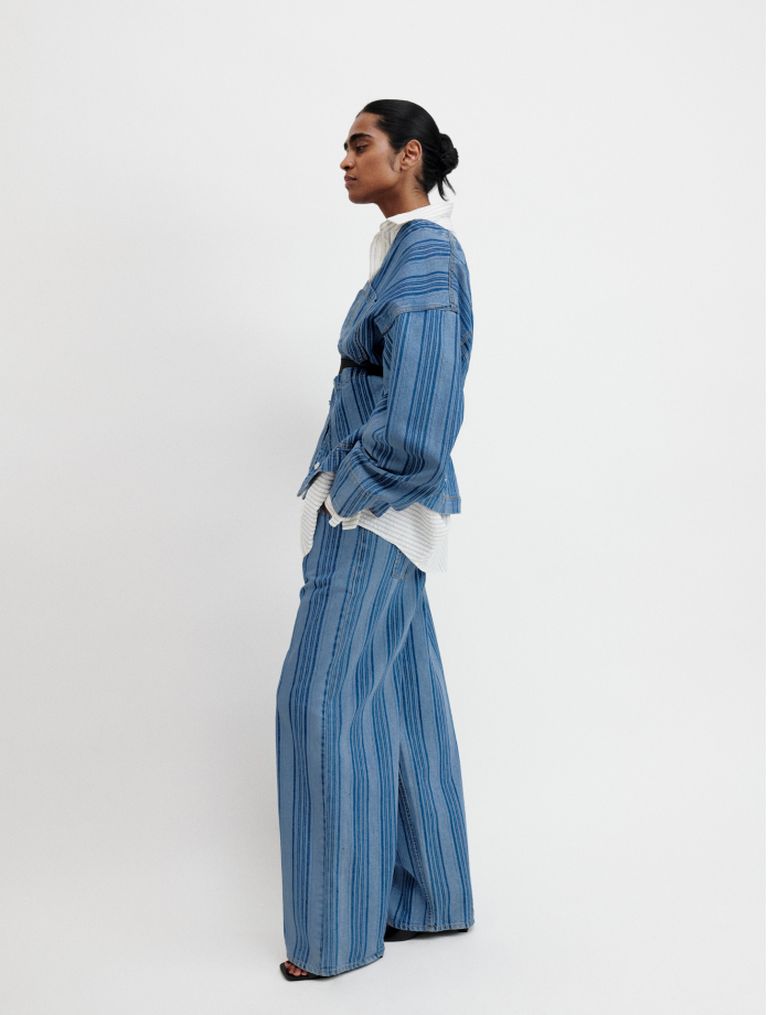 Person in a blue striped suit with wide-legged pants, white shirt, and sandals, standing against a plain white background.