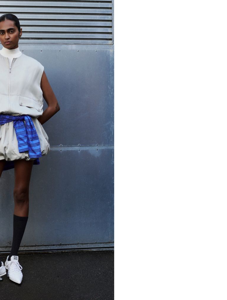 Model in a white sleeveless vest, blue skirt, knee-high socks, and sneakers, stands against a metal wall with a focused expression.