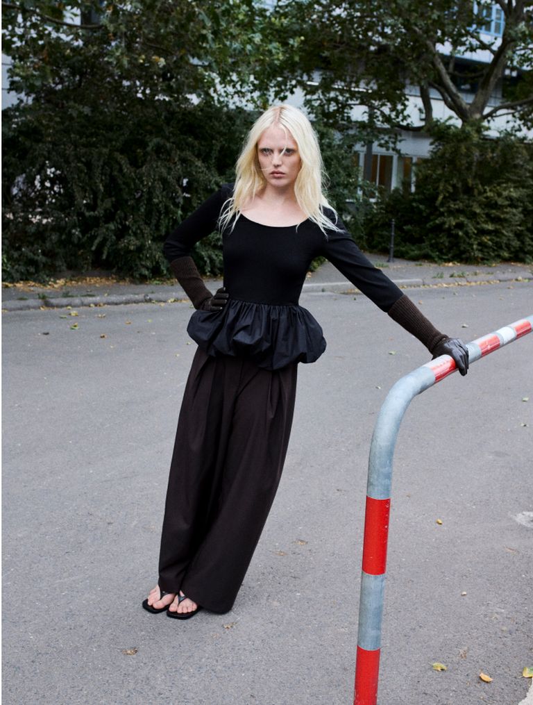 A person with long blonde hair wearing a black top and wide-leg pants leans against a street barrier, with trees and buildings in the background.