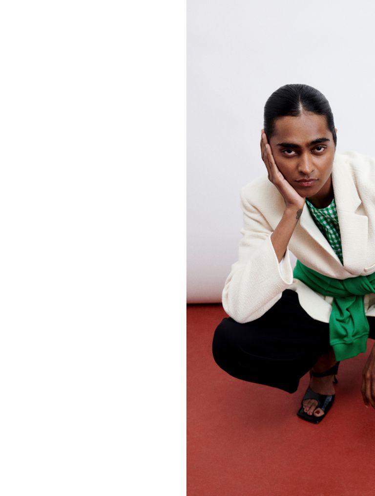 Person in a white blazer, green top, and black pants crouching on a red floor, resting their face on their hand, with a neutral expression.