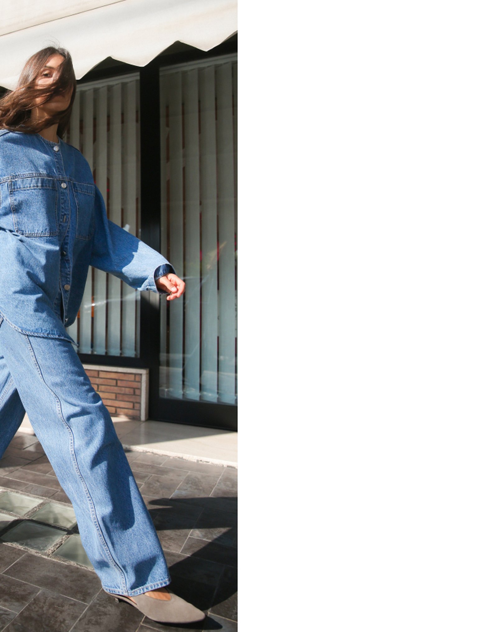 Person in a denim jacket and jeans walking on a tiled surface, with a building and vertical blinds in the background.