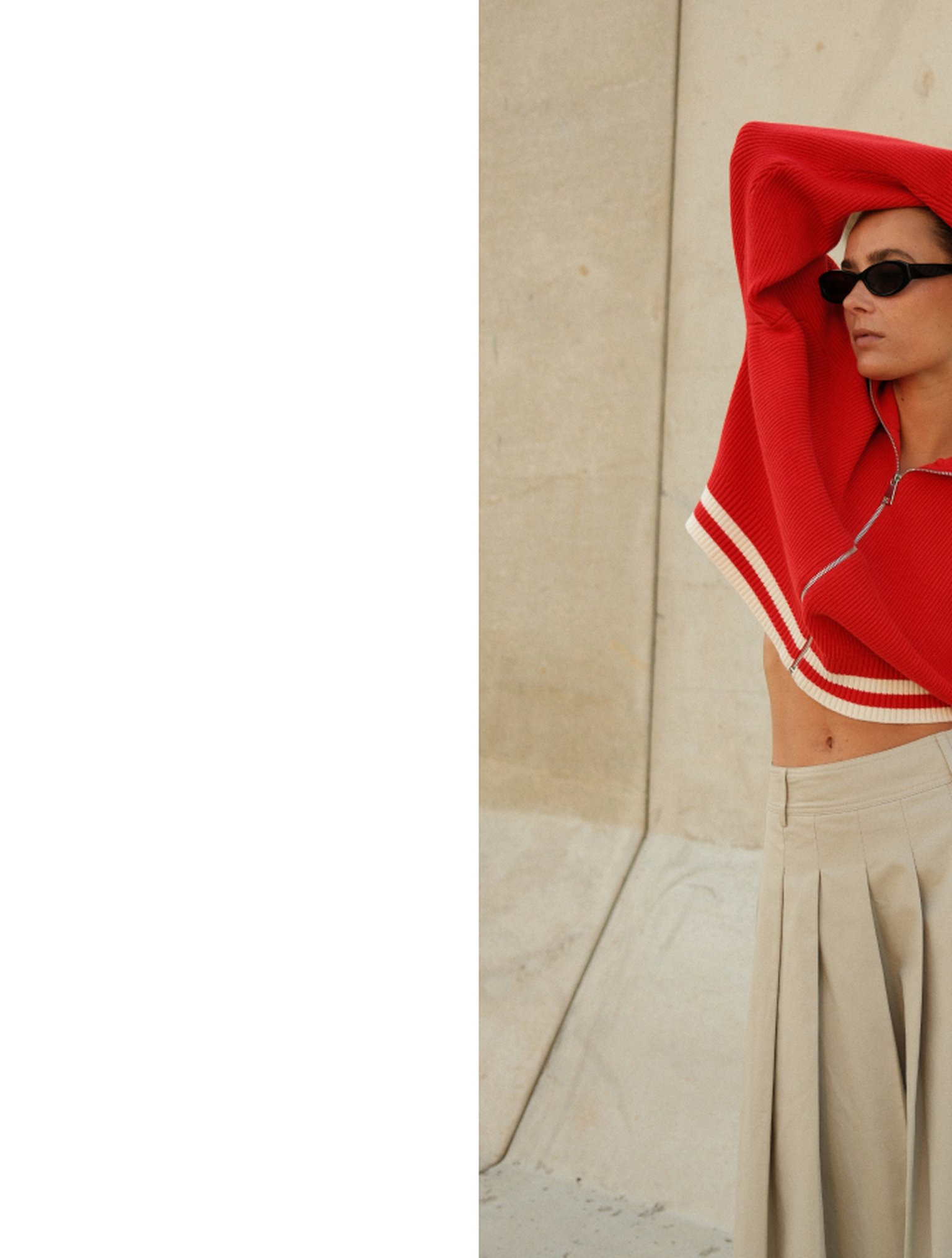 Woman wearing sunglasses with a red top partially over her head, standing against a beige wall in a pleated skirt.