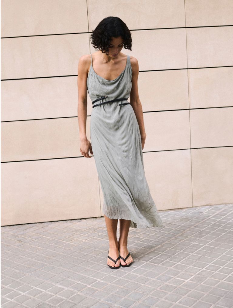 Person in a light green dress with black belt and sandals stands on a tiled pavement against a beige wall, looking down.