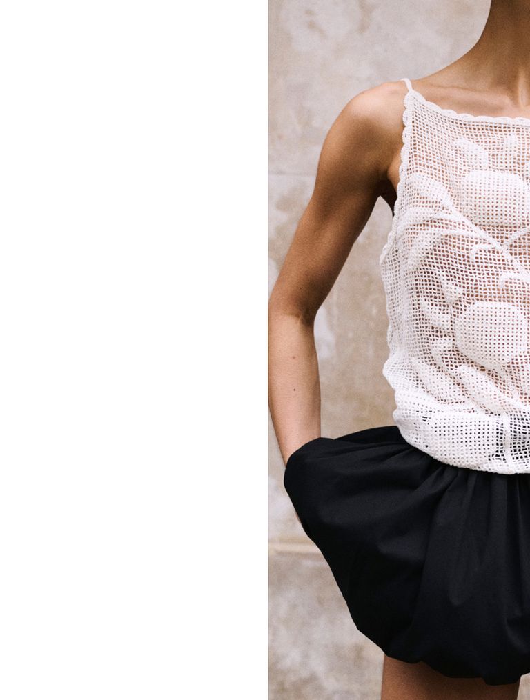 Person wearing a white crochet top and black skirt, standing against a textured beige wall, with arms slightly bent by their sides.