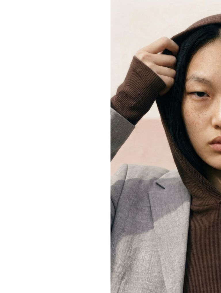 Person in a brown hood and gray blazer, partially covering their face with a hand, against a soft pink background.