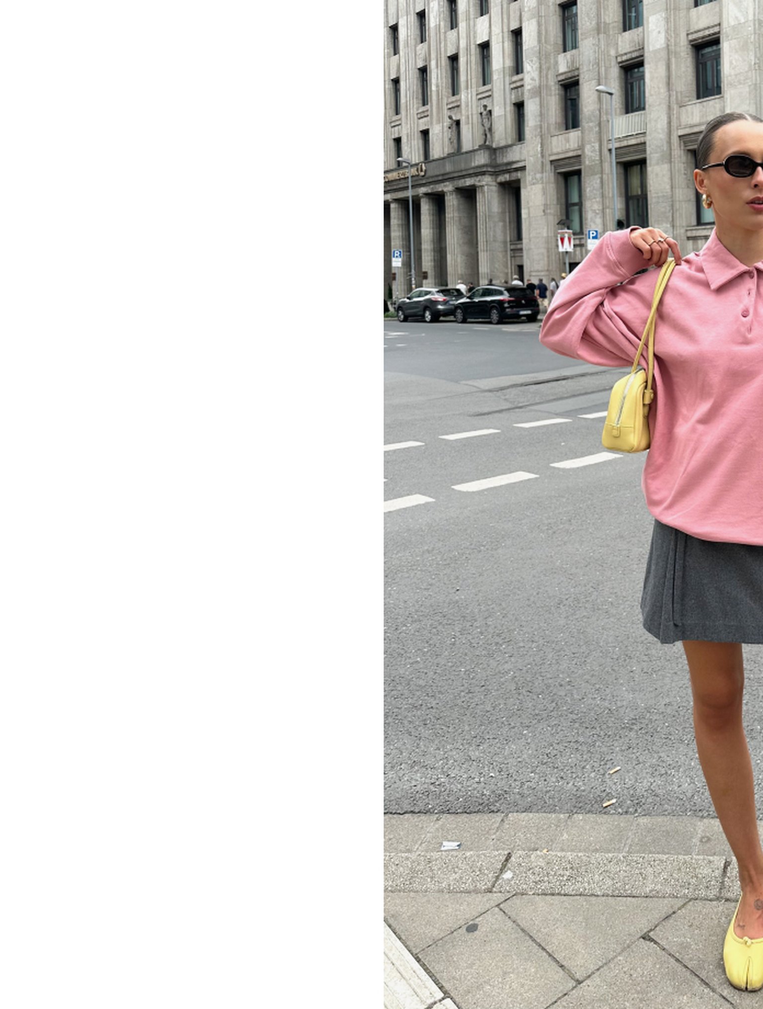 A person in a pink sweater and gray skirt stands at a city crosswalk, holding a yellow bag. Urban buildings are in the background.