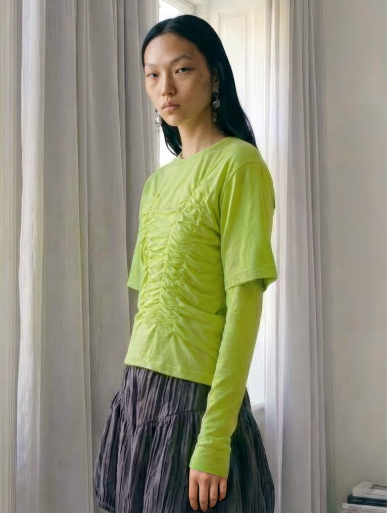A person wearing a bright green ruched top and a dark pleated skirt, standing in a room with soft light and curtains.
