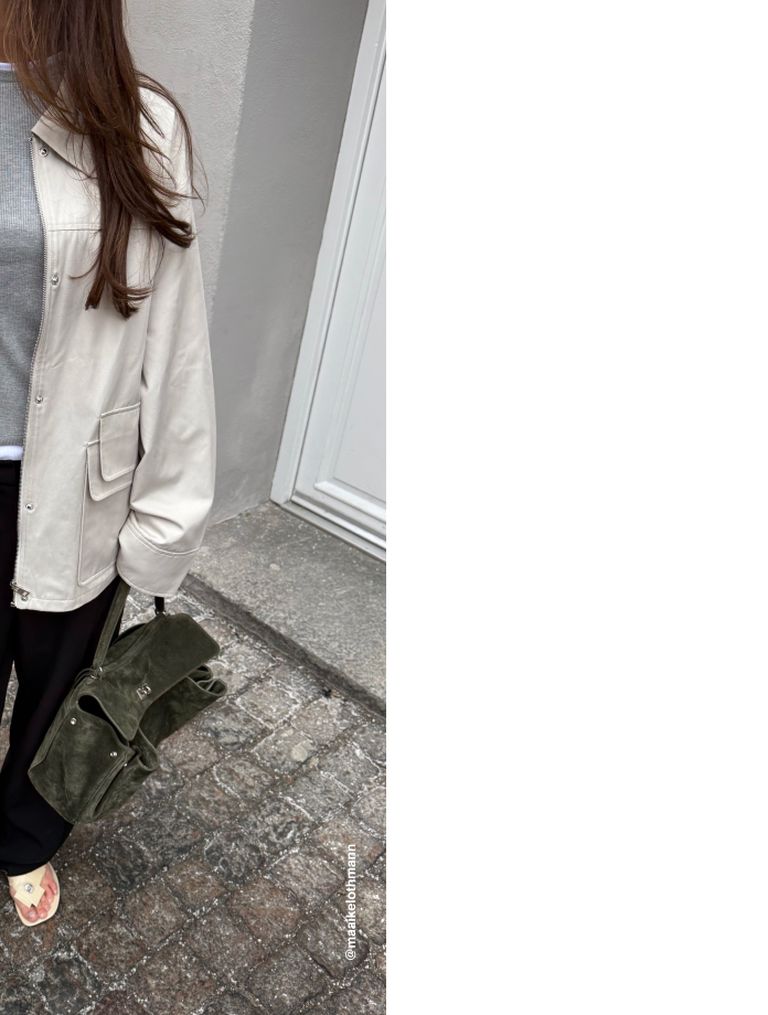 A person in a beige jacket and black pants holds an olive green suede bag. The background shows a gray wall and gray stone pavement.